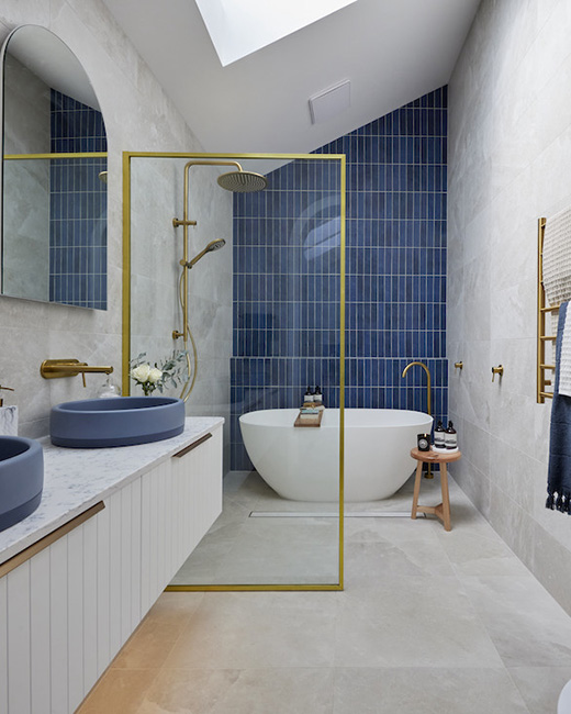 A modern bathroom showcasing blue tiled walls, a bathtub, a fixed shower panel with a brushed gold frame.